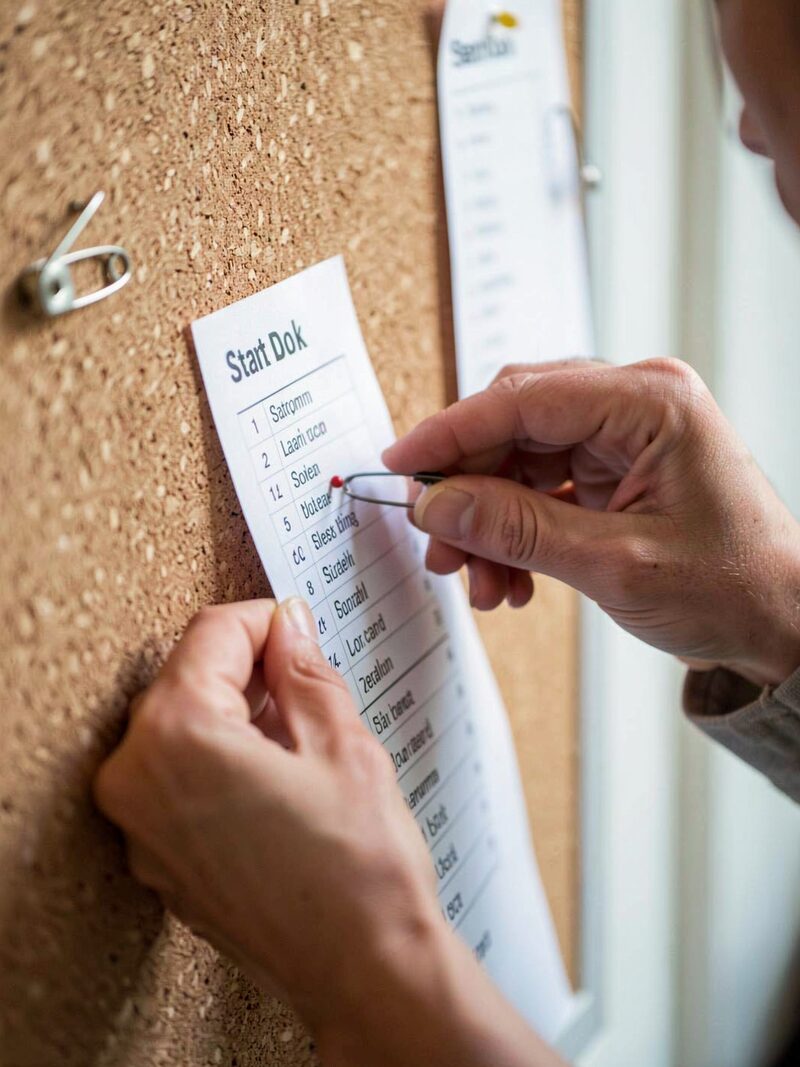A paper start sheet pinned to a folding table at a race registration desk in a parking lot at dawn, pens and blank waiver forms beside it, cyclists rolling past out of focus in the background