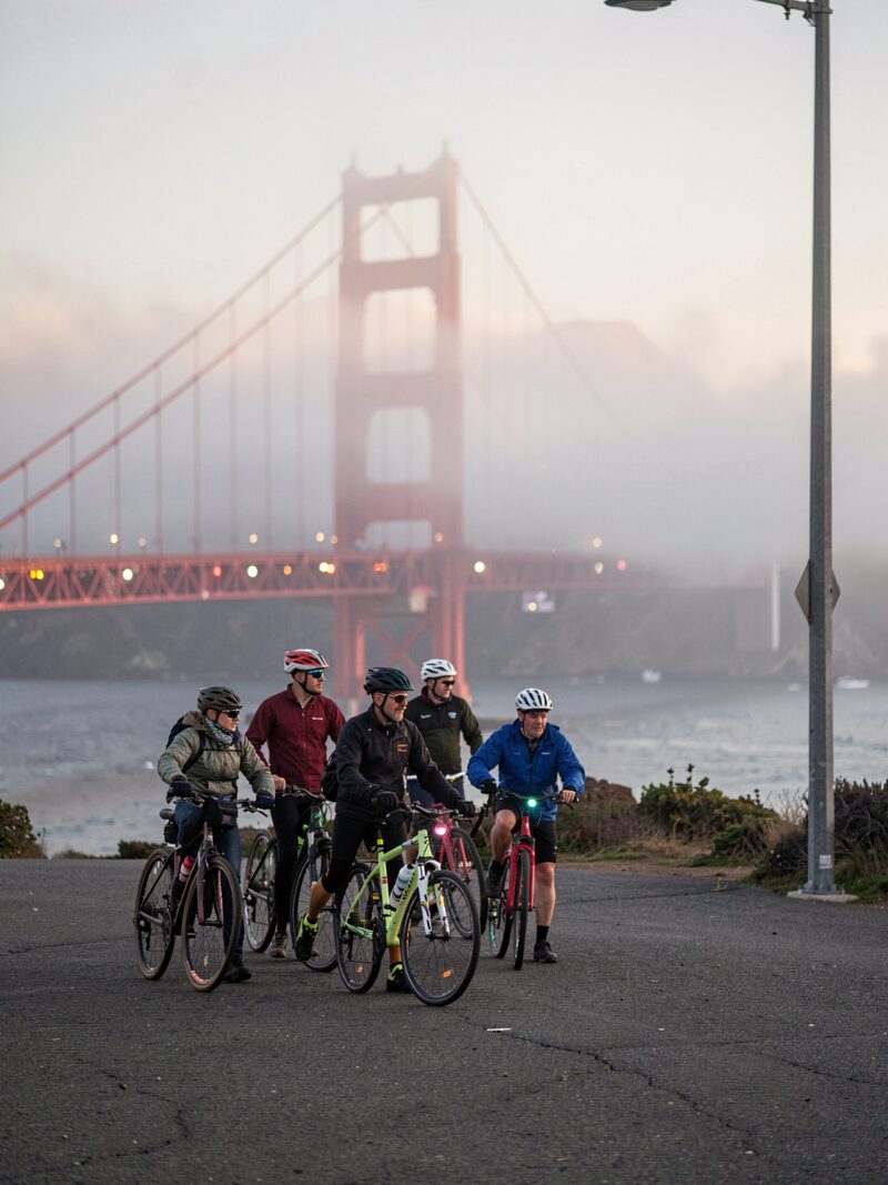A small group of cyclists gathering at the north end of the Golden Gate Bridge at dawn, red tower rising into fog, blinky lights and helmets