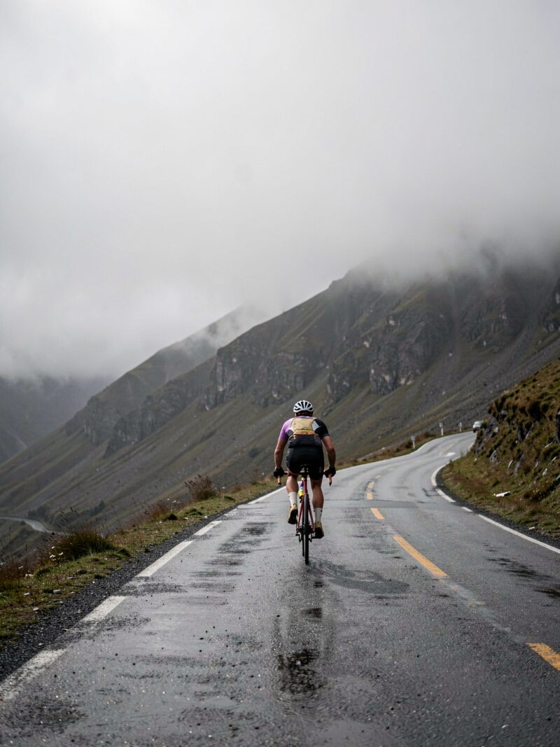 A lone cyclist climbing a steep wet mountain road into fog, low clouds over dark ridges, effort visible in the posture, photographed from behind
